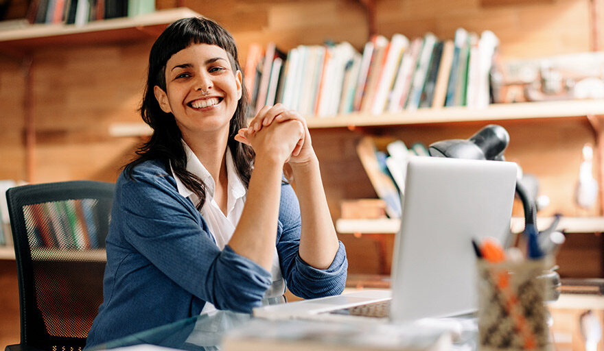 Woman sitting in front of laptop and smiling with bookcases behind her.