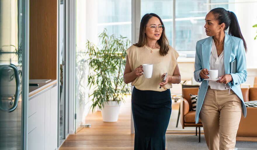 Two women in an office setting walking down the hallway together, holding coffee cups.