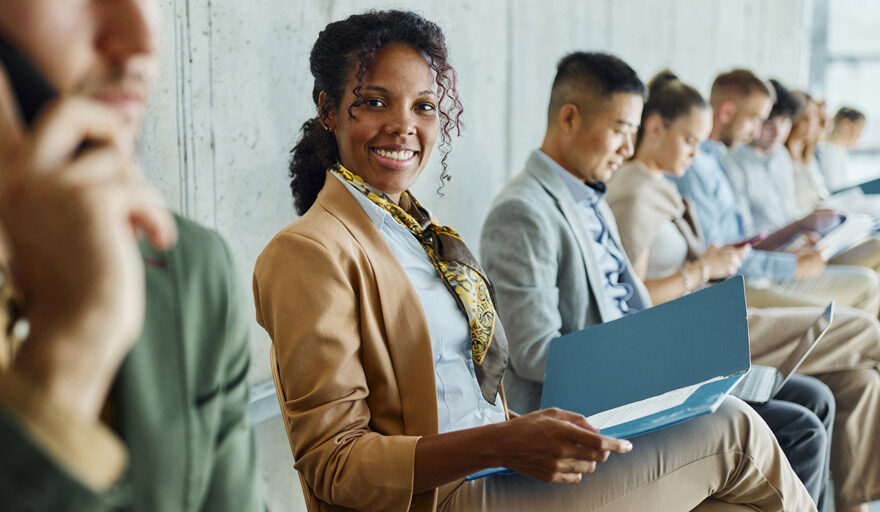 Group of happy, professional people sitting in chairs in a line against a wall.