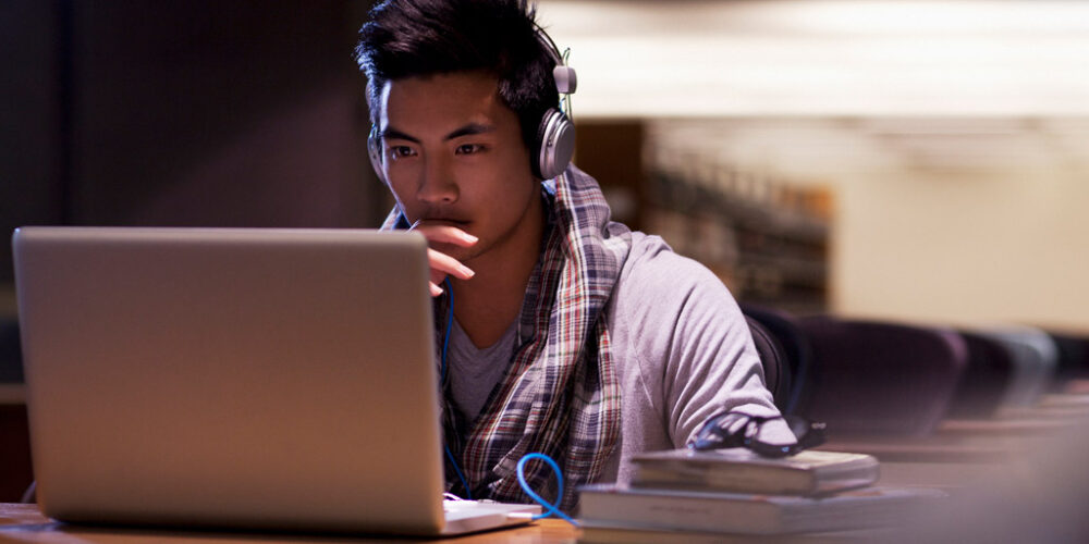 Young man wearing headphones looking at a laptop computer screen in a quiet library.