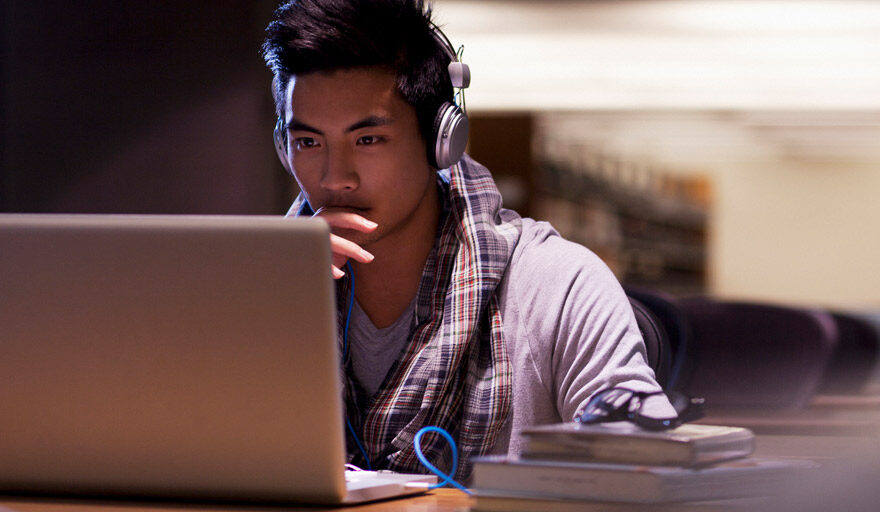 Young man wearing headphones looking at a laptop computer screen in a quiet library.
