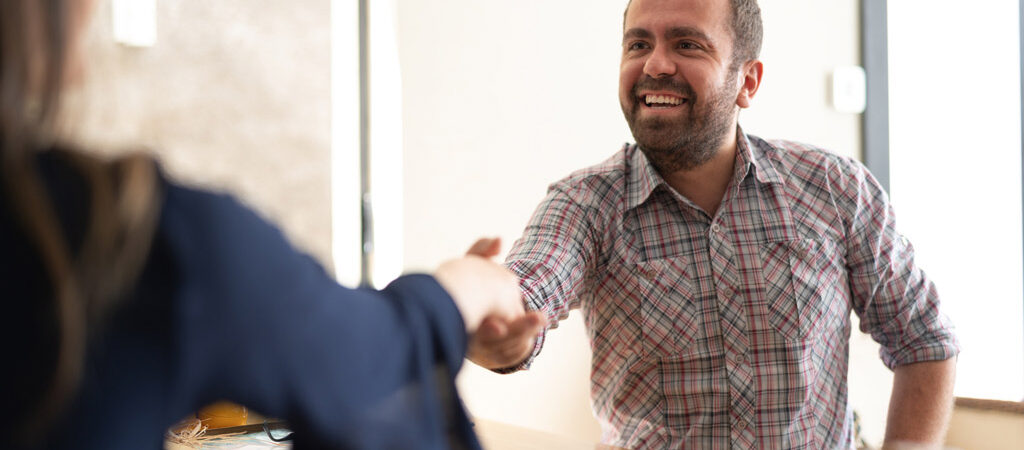 A smiling man reaches across the table to shake someone's hand.