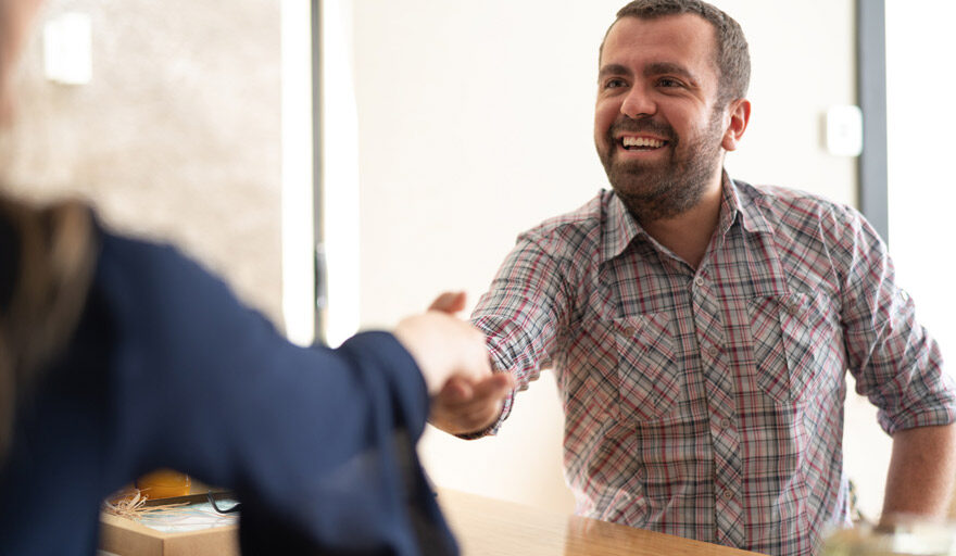 A smiling man reaches across the table to shake someone's hand.
