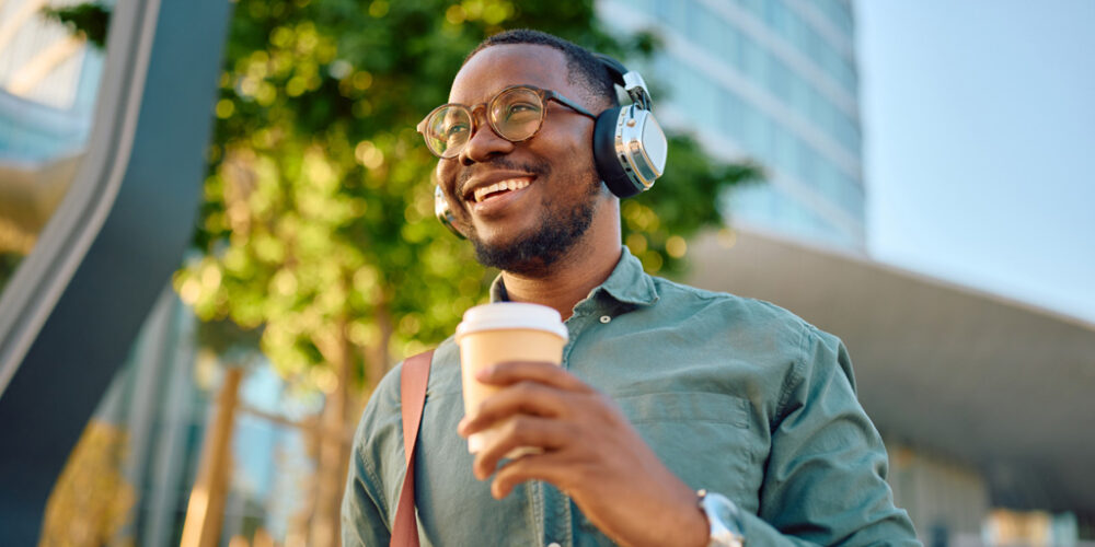 Smiling man wearing headphones and holding a to-go-cup, while walking outside.
