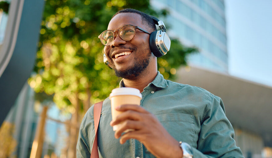 Smiling man wearing headphones and holding a to-go-cup outside.